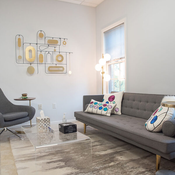 Modern psychotherapy office interior with gray couch, decorative pillows, accent chair, glass coffee table, and abstract wall art, reflecting a welcoming environment for therapy sessions.