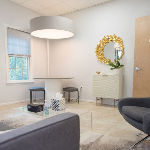 Therapy room interior featuring a round table, two chairs, a decorative mirror with golden leaves, a cabinet with a plant, and a comfortable gray chair, designed for child and family counseling sessions.