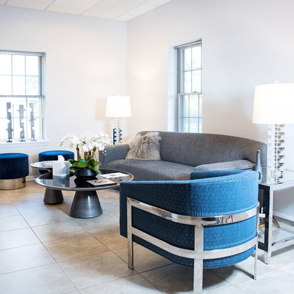 Modern therapy office interior with a gray sofa, blue accent chairs, a round coffee table, decorative plants, and warm lighting, reflecting a welcoming environment for counseling sessions.