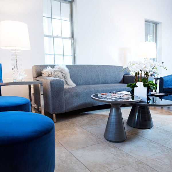 Modern therapy waiting room with gray sofa, blue accent chairs, glass coffee table, and floral arrangement, designed for comfort and relaxation in a mental health practice.