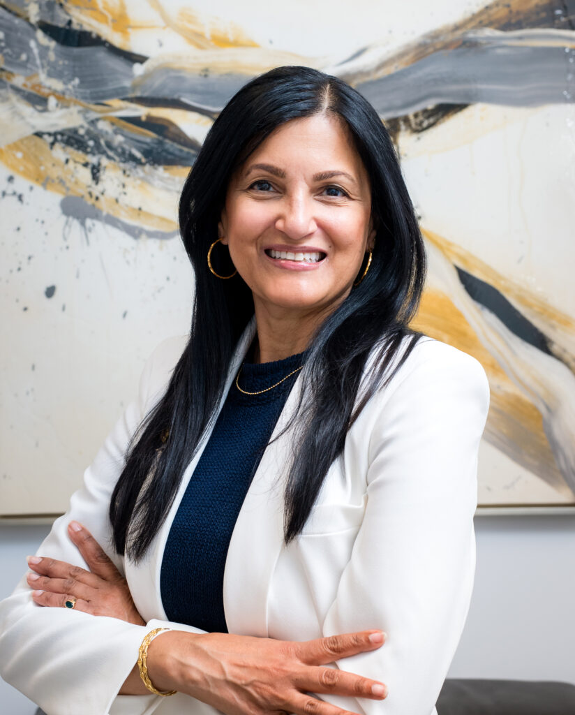 Hindi-speaking therapist smiling, dressed in a white blazer, with arms crossed, against an abstract background, representing Bergen County Therapists' counseling services for individuals and families.
