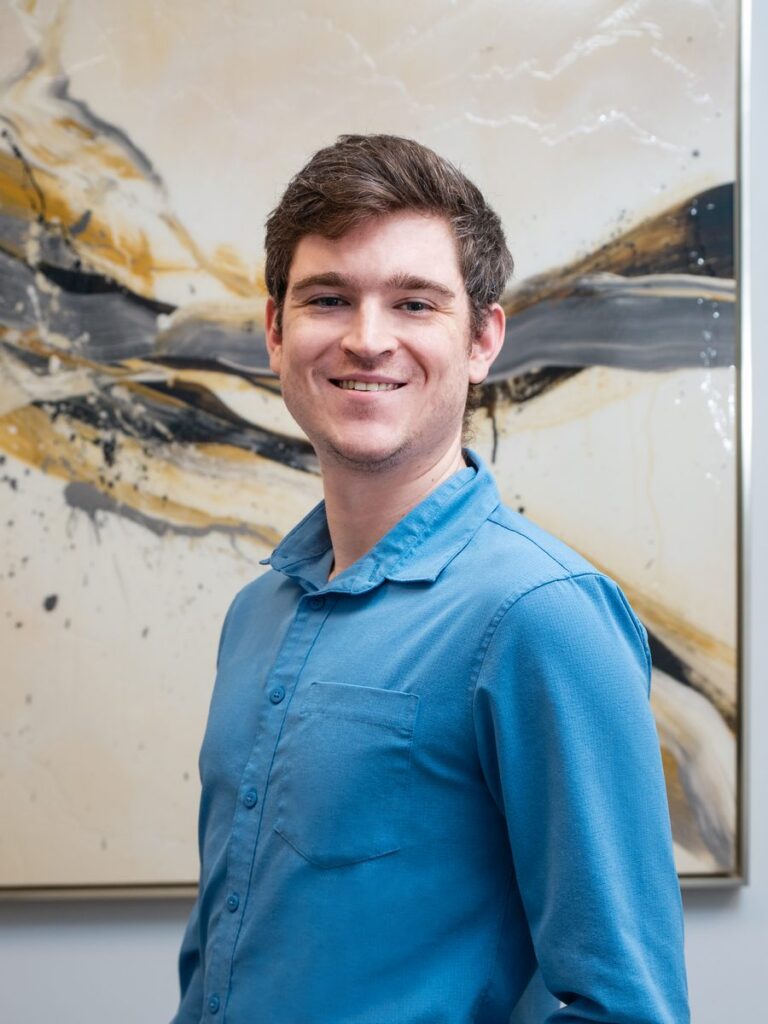 Liam Kane, therapist at Bergen County Therapists, smiling in a blue shirt against an abstract art background, representing mental health services including counseling and therapy.