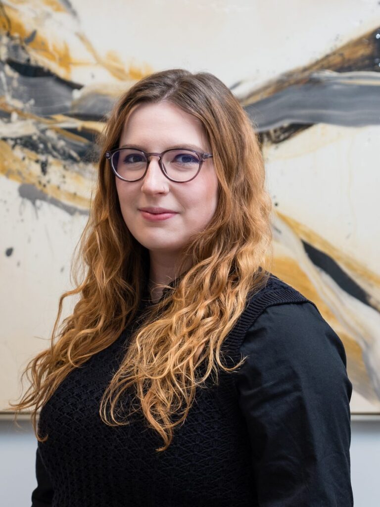 Tierney Byrne, Counseling Intern, with long curly hair and glasses, standing in front of an abstract art background, representing mental health services for adults, teens, and children, specializing in anxiety, depression, and stress management.