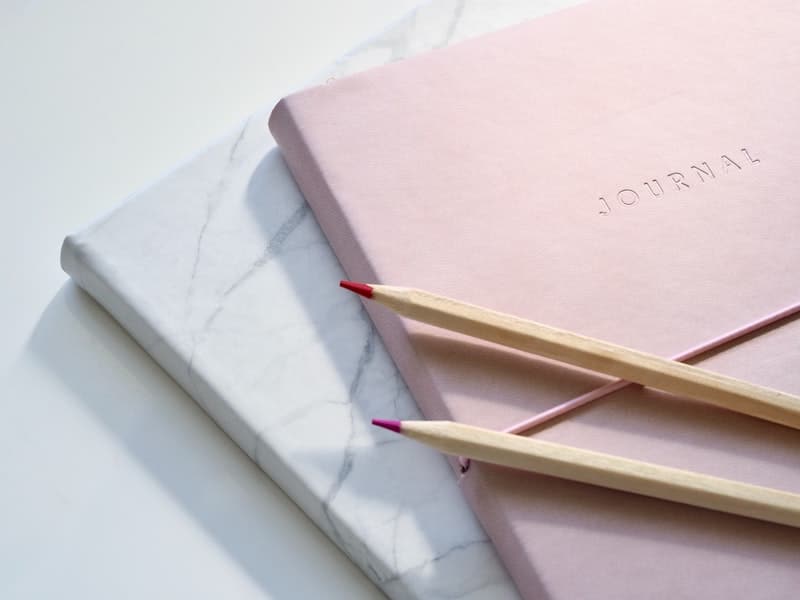 Pink journal and white marble notebook with colored pencils, representing thoughtful gift ideas for mental health during the holiday season.