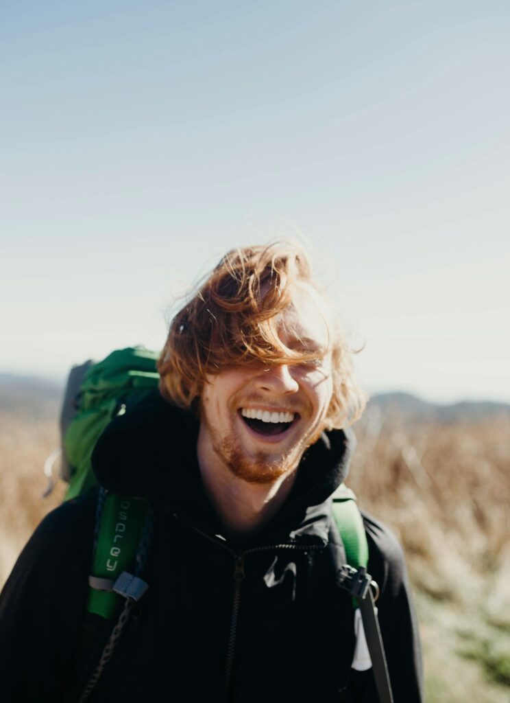 Smiling young man with tousled hair and a backpack, embodying joy and well-being in a natural outdoor setting, reflecting themes of self-care and mental health.