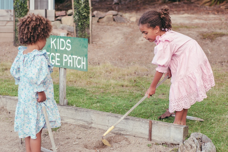 Two children gardening in a patch labeled "KIDS GARDEN PATCH," one digging with a tool and the other observing, showcasing playful engagement with nature.