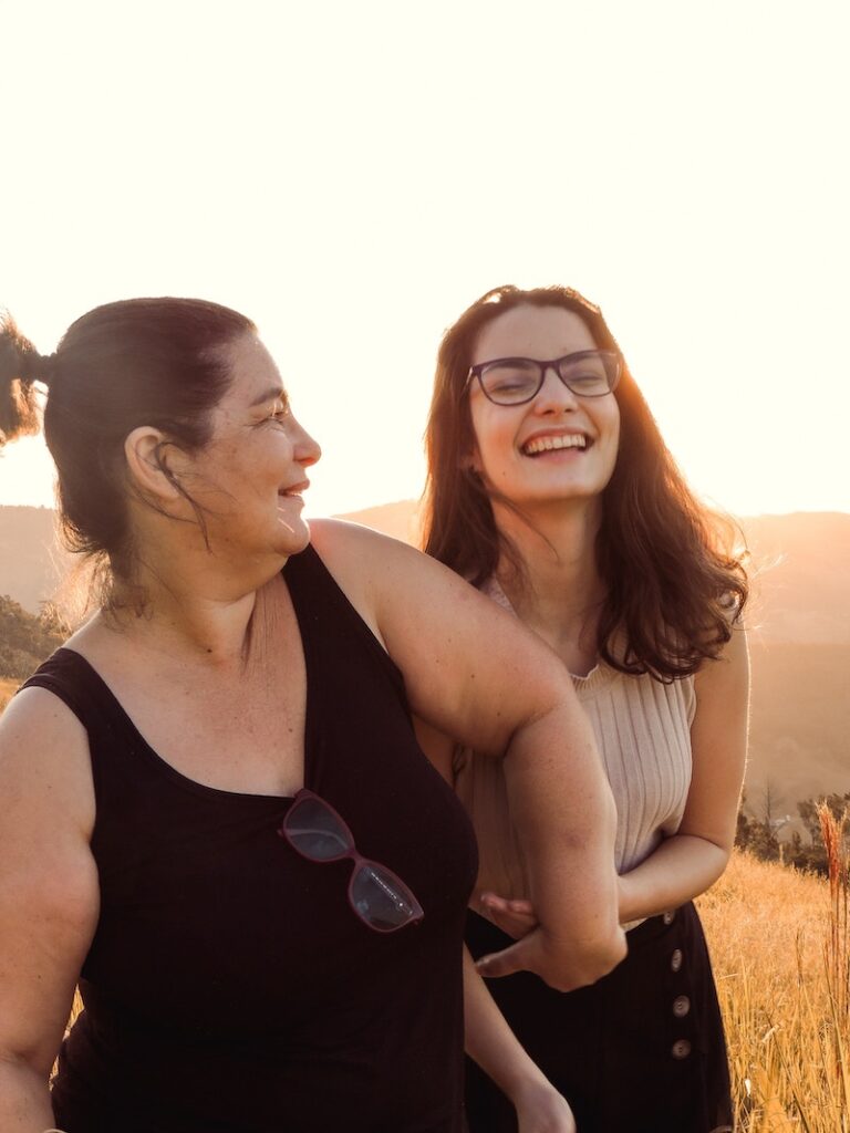 Two women smiling and enjoying a moment together in a sunlit outdoor setting, representing the joy of parenting and family connections.