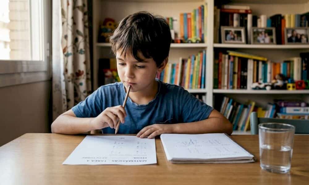 Ni&ntilde;o pensando en mesa de comedor, realizando tareas escolares, con hojas de ejercicios y vaso de agua, en un entorno familiar que refleja la importancia del apoyo emocional en la ansiedad infantil.