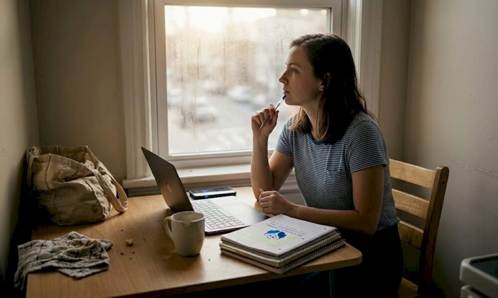 Person reflecting at a kitchen table workspace with a laptop, notebooks, and a coffee mug, contemplating the effects of minority stress on mental health.