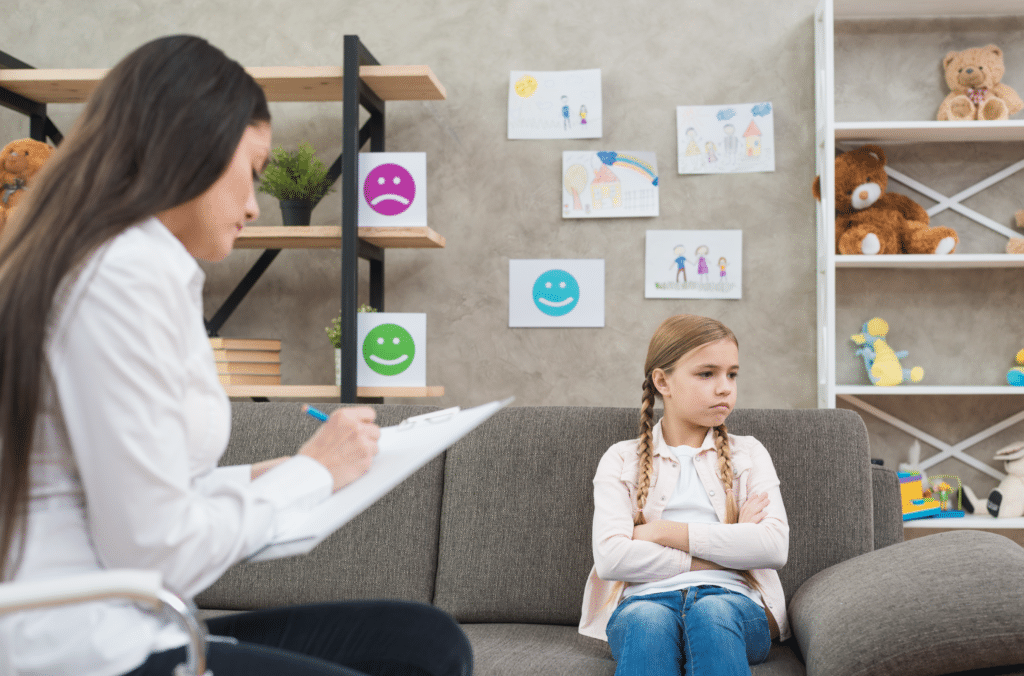 Child therapist taking notes while a young girl sits on a couch with crossed arms in a Paramus, New Jersey playroom office