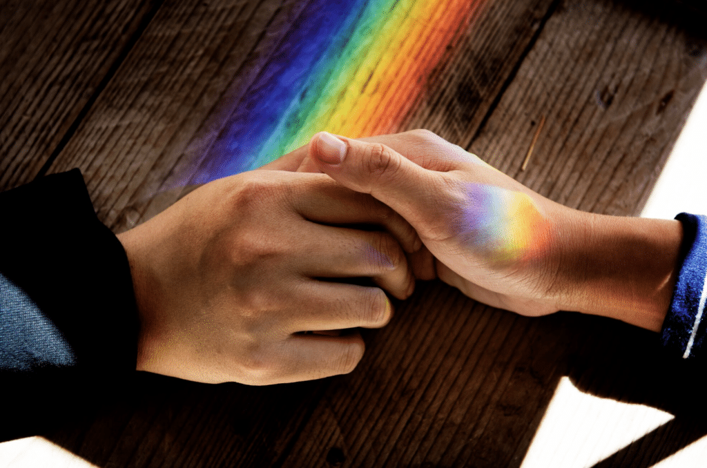 Two people holding hands with a rainbow light across a wooden table, representing LGBTQ therapy in Paramus, New Jersey